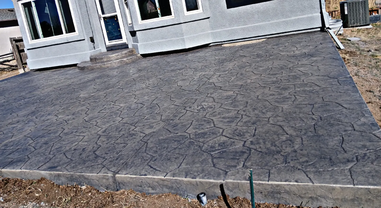 A wide charcoal stone-pattern stamped concrete patio behind a two-story house, cut into panels with control joints.