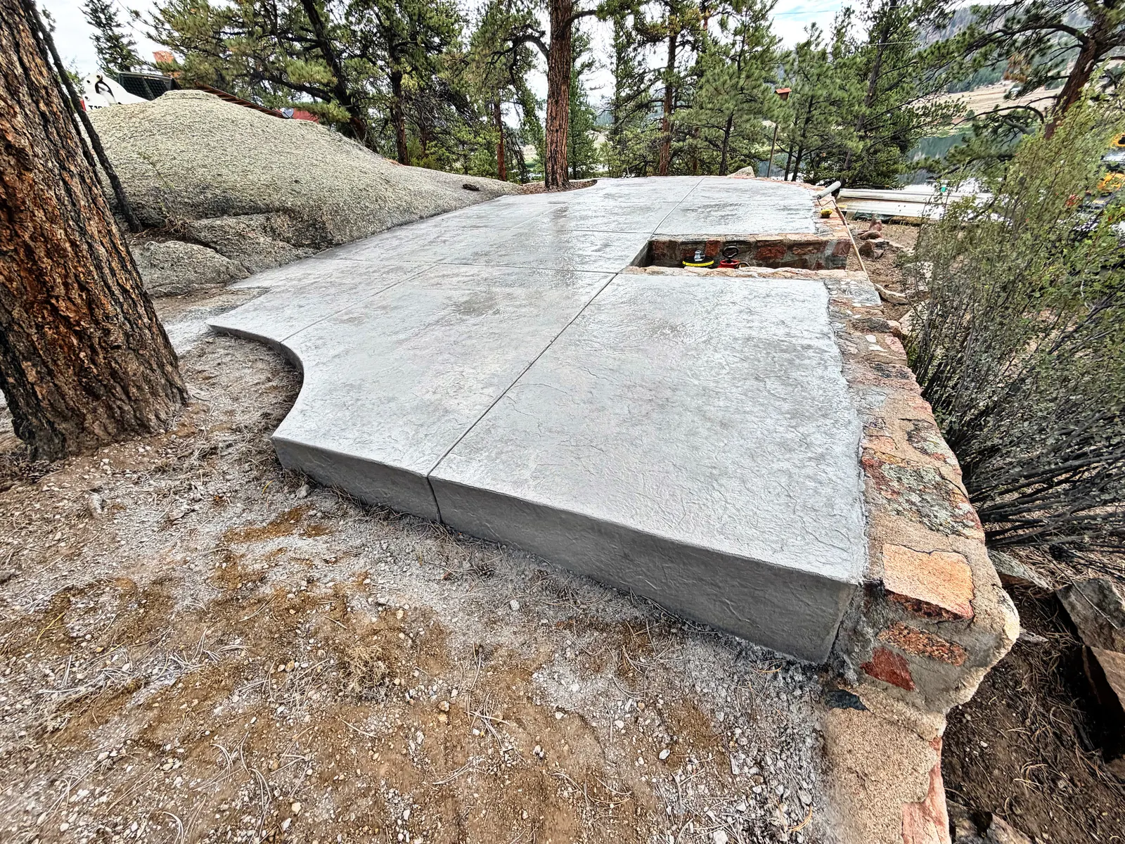 A raised grey textured concrete slab with a curved edge around a ponderosa pine, beside a granite outcrop.