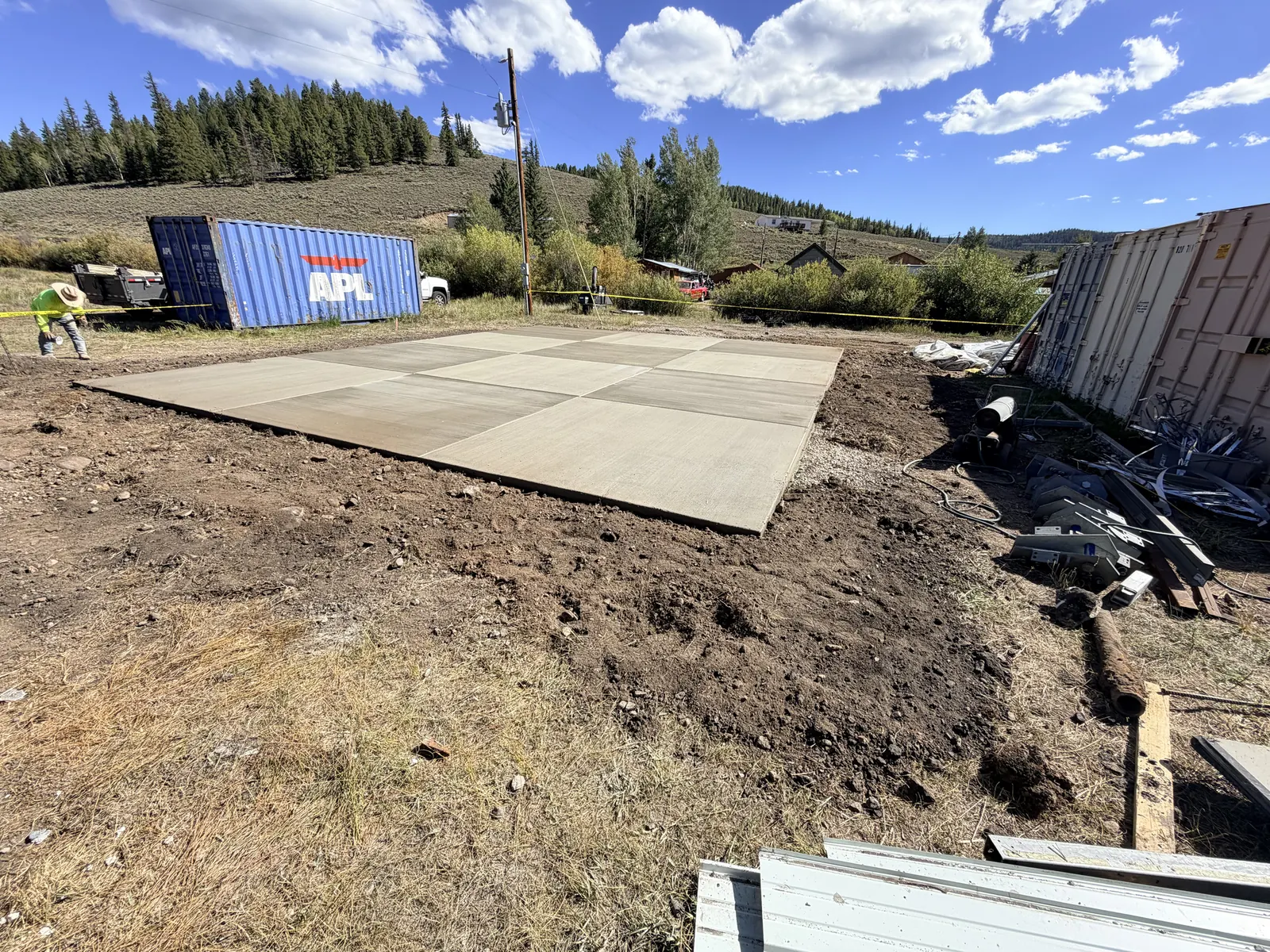 A large new concrete slab with saw-cut joints on a cleared mountain lot under a blue sky.