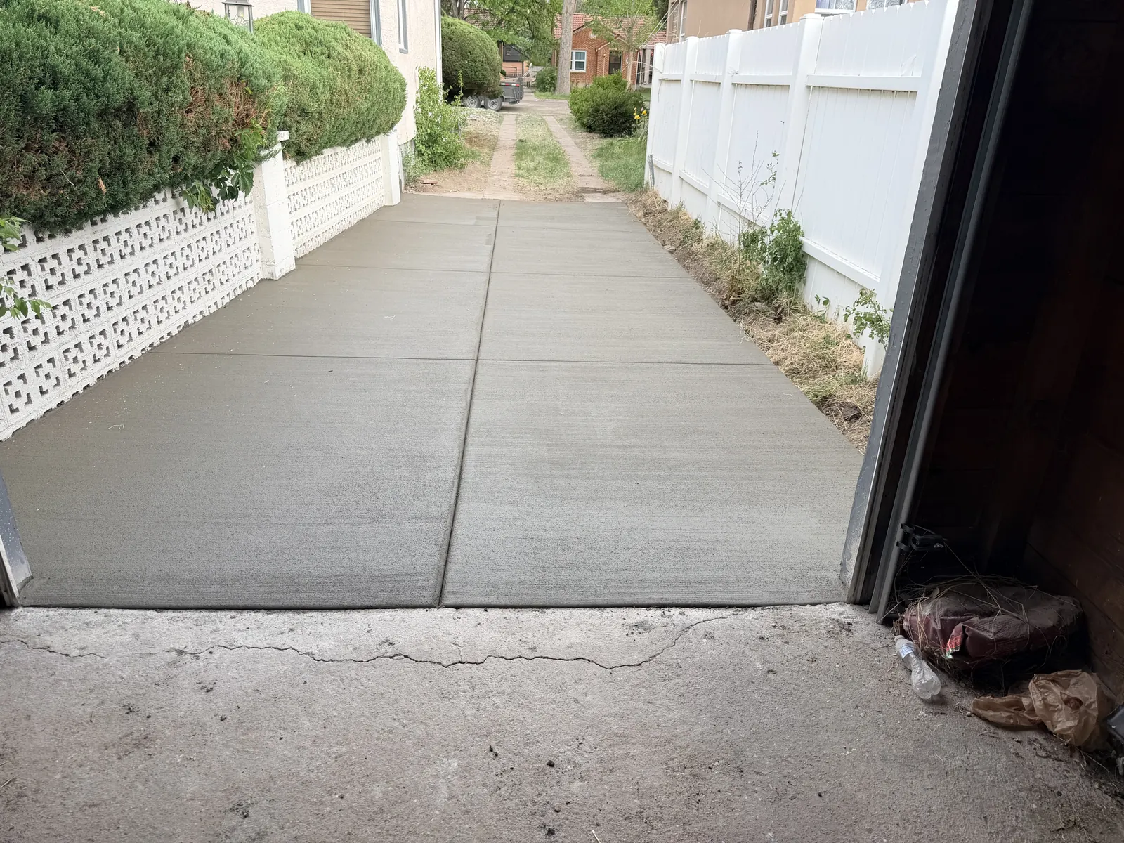 A long broom-finished concrete slab running from a garage door down a side yard between a fence and a hedge.